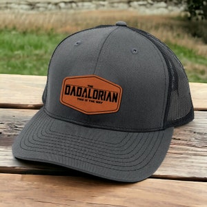 May include: A gray and black trucker hat with a brown leather patch that says "The Dadalorian This is the Way".