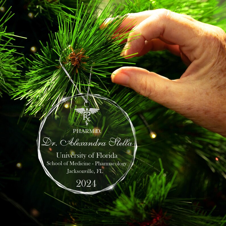 K&ouml;nnte beinhalten: Ein transparenter Weihnachtsbaumschmuck aus Glas mit einer silbernen Inschrift. Die Inschrift lautet "PHARM.D. Dr. Alexandra Stella University of Florida School of Medicine - Pharmacology Jacksonville, FL 2024".