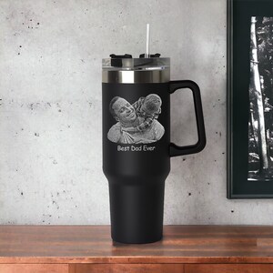 May include: Black stainless steel travel mug with a black handle. The mug has a photo of a father and child engraved on the front with the text "Best Dad Ever".