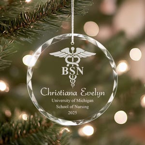 May include: A clear glass Christmas ornament with a silver ribbon. The ornament is engraved with a caduceus symbol, the letters "BSN", the name "Christiana Evelyn", "University of Michigan School of Nursing", and the year "2025".