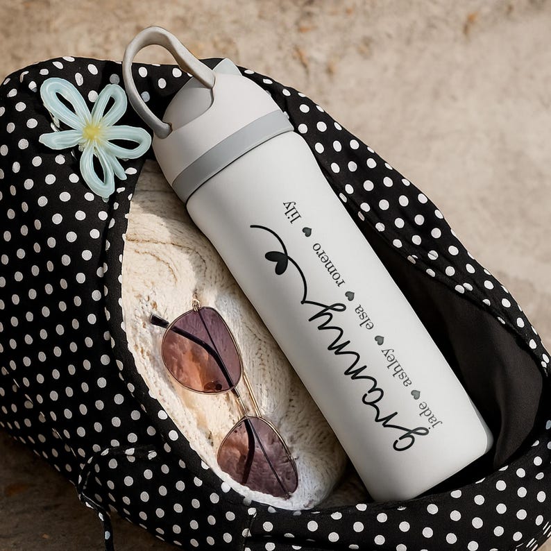 May include: A white water bottle with the word "Granny" in cursive, with names like "Jade" and "Lily" listed below. The bottle has a gray handle and a black and white polka dot bag is in the background. Sunglasses are also visible.