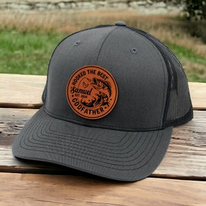May include: A gray and black trucker hat with a brown leather patch that says "Hooked the Best Samuel Est. 2024 Godfather" with a fish jumping out of the water.