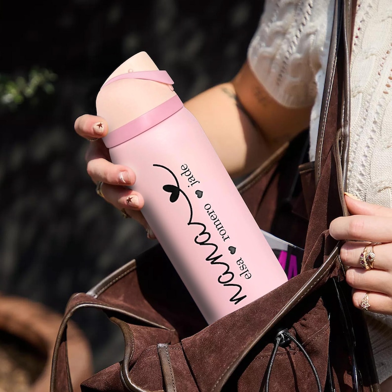 May include: A pink insulated water bottle with a light pink lid and strap. The bottle has the name "Nana" in a cursive font, along with the names "Elsa Romero Jade" in a smaller font. The bottle is being held.