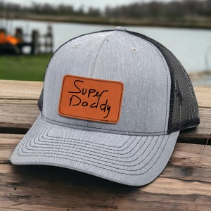 May include: A gray trucker hat with a black mesh back and a curved brim. The hat features a brown leather patch with the handwritten text "Super Daddy". The hat is sitting on a wooden surface.
