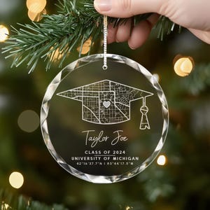 May include: A clear glass Christmas ornament with a white outline of a graduation cap and a map of the University of Michigan. The ornament also includes the text "Taylor Joe, Class of 2024, University of Michigan, 42°16'37.7"N 83°44'17.5"W".