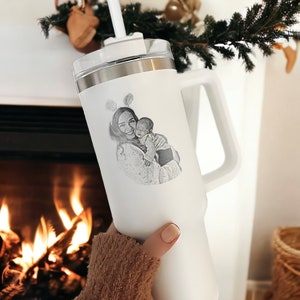 May include: White travel mug with a handle and a black and white photo of a mother and child engraved on the side.