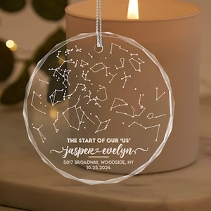 May include: Clear, faceted glass ornament with a constellation design. The text "THE START OF OUR 'US'" is above the names "jasper & evelyn" and a date. A silver string is attached for hanging. The ornament is near a lit candle.