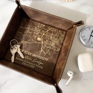 May include: A brown leather catchall tray with a gold engraved map of New York University. The tray has the text "New York University, Christina Evelyn, Bachelor of Science in Nursing, Class of 2025".
