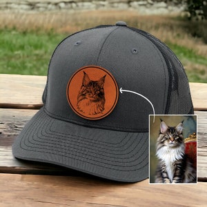 May include: A gray and black trucker hat with a brown leather patch featuring a black and white image of a cat's face.