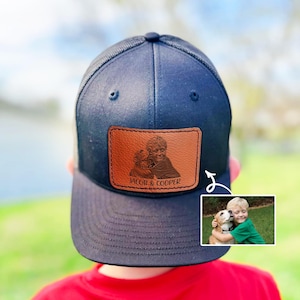 May include: A black and brown trucker hat with a personalized leather patch featuring a photo of a boy and his dog. The patch has the text "Jacob & Cooper" on it.