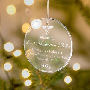 K&ouml;nnte beinhalten: Ein transparenter Weihnachtsbaumschmuck aus Glas mit einer silbernen Inschrift. Der Schmuck zeigt ein medizinisches Symbol, den Text "PHARM.D.", "Dr. Alexandra Stella", "University of Florida", "School of Medicine - Pharmacology", "Jacksonville, FL" und "2024".