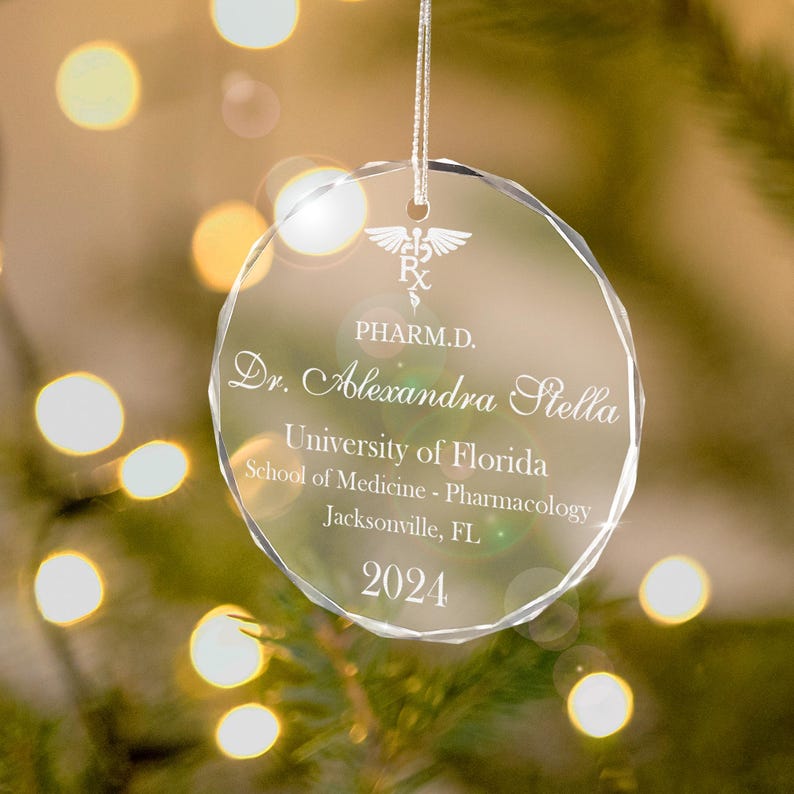 K&ouml;nnte beinhalten: Ein transparenter Weihnachtsbaumschmuck aus Glas mit einer silbernen Inschrift. Der Schmuck zeigt ein medizinisches Symbol, den Text "PHARM.D.", "Dr. Alexandra Stella", "University of Florida", "School of Medicine - Pharmacology", "Jacksonville, FL" und "2024".