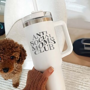 以下が含まれることがあります： 黒の取っ手と「Anti Social Moms Club」という白黒のグラフィックが描かれた白いステンレス製のトラベルマグです。