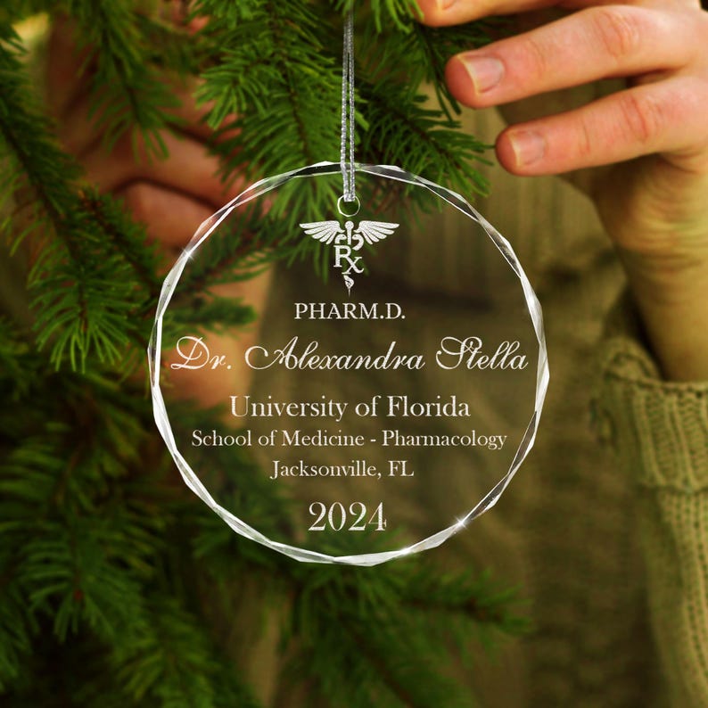 K&ouml;nnte beinhalten: Ein transparenter Weihnachtsbaumschmuck aus Glas mit einer silbernen Inschrift. Die Inschrift lautet "PHARM.D. Dr. Alexandra Stella University of Florida School of Medicine - Pharmacology Jacksonville, FL 2024".