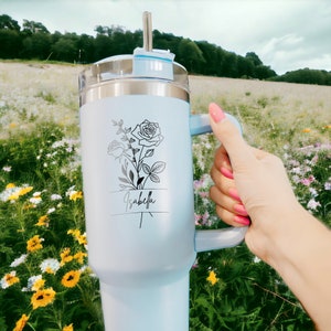May include: A light blue stainless steel travel mug with a black floral design and the name "Isabella" on it. The mug has a handle and a lid with a straw.