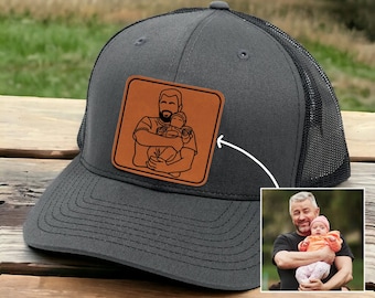 May include: A black and gray trucker hat with a brown leather patch featuring a line drawing of a person holding two children. The patch is on the front of the hat.