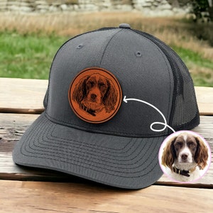 May include: A gray and black trucker hat with a brown leather patch featuring a black and white illustration of a dog's head. The patch is attached to the front of the hat.