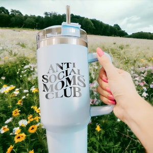 以下が含まれることがあります： 黒のハンドルと「Anti Social Moms Club」という黒と白のグラフィックが描かれた、薄い青色のステンレス製トラベルマグです。