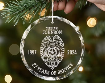 Könnte beinhalten: Ein transparenter Weihnachtsbaumschmuck aus Glas mit einem silbernen Polizeibadge-Design. Der Schmuck zeigt den Text "Sergeant Johnson", "Serve and Protect", "Police Officer", "Police", "1997", "2024", "1328" und "27 Years of Service".