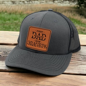 May include: A gray and black trucker hat with a brown leather patch that reads "First Dad - Now Grandpa" with the names Hannah and David and the names of the grandchildren.
