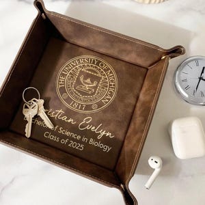 May include: A brown leather valet tray with a gold University of Michigan logo and the inscription "Christian Evelyn Bachelor of Science in Biology Class of 2025".