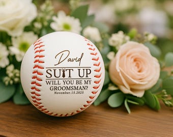 May include: A white baseball with red stitching and the text "David SUIT UP WILL YOU BE MY GROOMSMAN? November.15.2025". The baseball is surrounded by white and peach-colored flowers and greenery, resting on a wooden surface.