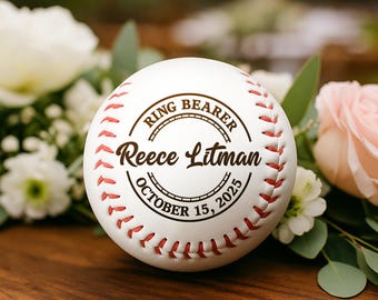 May include: A white baseball with red stitching and the text "RING BEARER" above "Reece Litman" and "OCTOBER 15, 2025" in a circular design. The baseball is surrounded by white and pink flowers on a wooden surface.