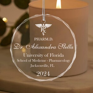 K&ouml;nnte beinhalten: Ein transparenter Weihnachtsbaumschmuck aus Glas mit einer wei&szlig;en Aufschrift. Der Schmuck zeigt ein medizinisches Symbol, den Text "PHARM.D.", "Dr. Alexandra Stella", "University of Florida", "School of Medicine - Pharmacology", "Jacksonville, FL" und das Jahr "2024".