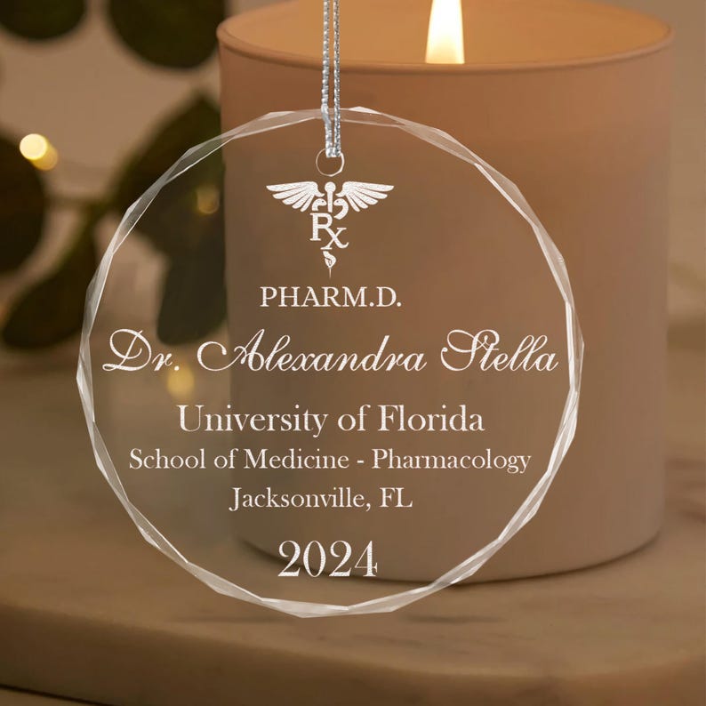 K&ouml;nnte beinhalten: Ein transparenter Weihnachtsbaumschmuck aus Glas mit einer wei&szlig;en Aufschrift. Der Schmuck zeigt ein medizinisches Symbol, den Text "PHARM.D.", "Dr. Alexandra Stella", "University of Florida", "School of Medicine - Pharmacology", "Jacksonville, FL" und das Jahr "2024".