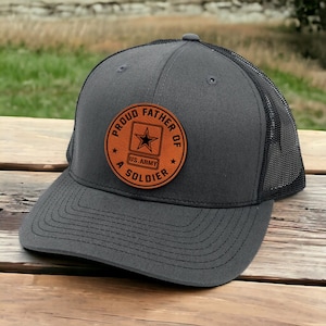 May include: A charcoal gray trucker hat with a black mesh back. The hat features a brown leather patch with the words "PROUD FATHER OF A SOLDIER" and a US Army emblem.