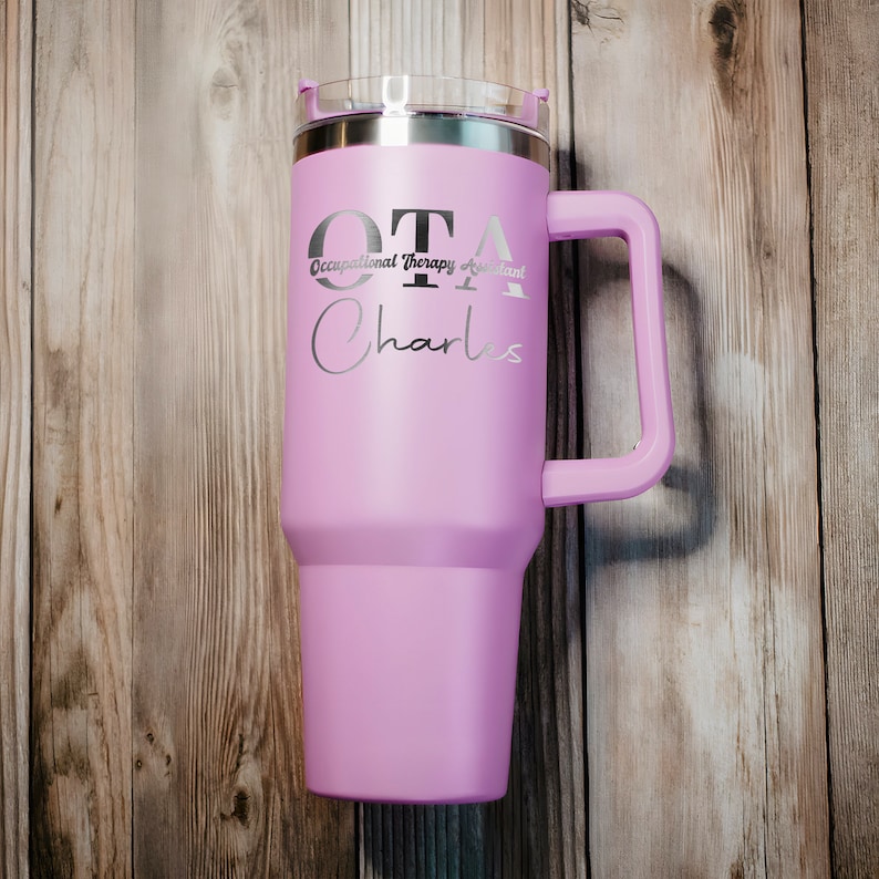 Pu&ograve; includere: Una tazza da viaggio in acciaio inossidabile rosa con un manico nero. La tazza ha il testo "OTA Occupational Therapy Assistant Charles" inciso sul lato.
