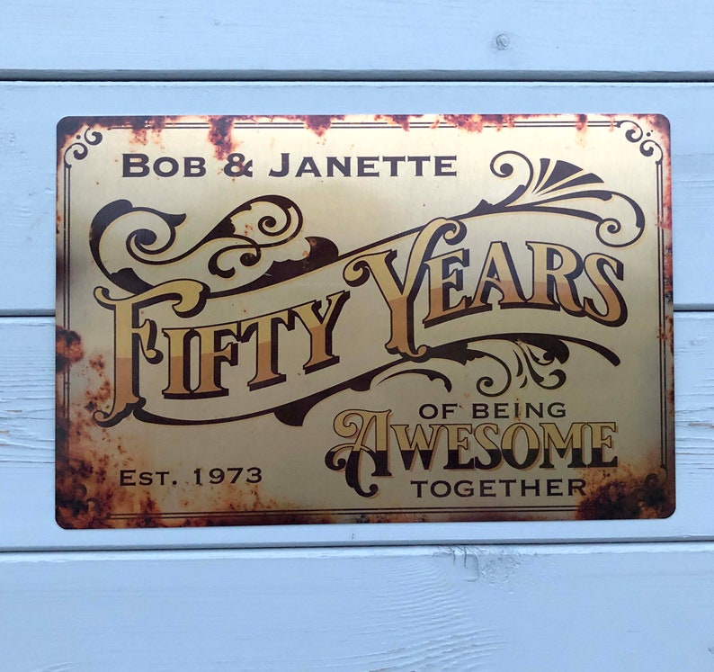 May include: A gold metal sign with a rusty finish. The sign reads "Bob & Janette Fifty Years of Being Awesome Together Est. 1973".
