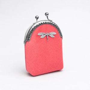 Pode incluir: Uma carteira de moedas cor-de-rosa com um fecho de metal prateado e um adorno de libélula. A carteira tem um topo arredondado e um corpo retangular. O fecho apresenta dois pequenos botões decorativos.