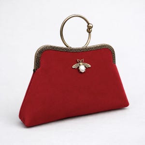 May include: A deep red handbag with a smooth texture, featuring an antique brass-toned metal frame with intricate filigree detailing. A circular brass handle sits atop the frame, and a central bee-shaped ornament with a white pearl body is attached.