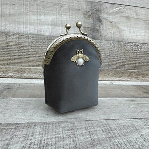 May include: A gray velvet coin purse with a brass clasp and a decorative bee emblem. The purse has a rounded top and a rectangular base. The clasp features two small, round brass accents.