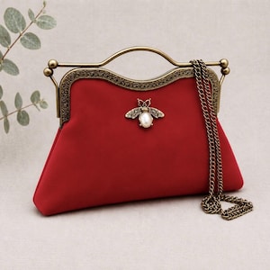 May include: A deep red handbag featuring a brass-toned metal frame with an arched handle and a detachable chain strap. The front is adorned with a central bee-shaped embellishment that has a white pearl-like body and intricate metal wings.