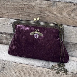 May include: A purple velvet clutch bag with a gold chain strap and a bee-shaped clasp with a purple gemstone.