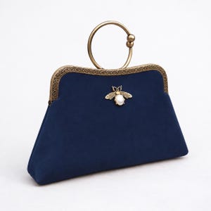 May include: A navy blue suede clutch bag featuring an ornate antique brass frame and a circular handle. The front is decorated with a central bee emblem crafted from brass and a white pearl-like stone, adding a distinct detail to the design.