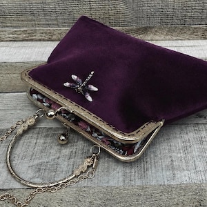May include: A deep purple velvet clutch bag featuring a gold-toned metal frame with a kiss-lock closure. A silver-toned dragonfly embellishment is attached to the front. The bag includes a circular gold metal handle and a detachable gold chain strap.