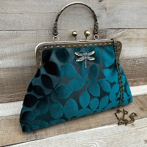 May include: A teal blue velvet clutch bag with a gold metal frame and a dragonfly brooch. The bag has a chain strap and a top handle.
