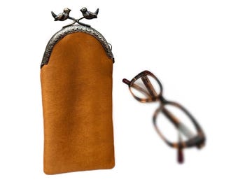 Może przedstawiać: Brązowe etui na okulary z ozdobnym metalowym zapięciem z dwoma ptakami. Obok etui znajduje się para okularów w kolorze skorupy żółwia. Etui wykonane jest z teksturowanego materiału.