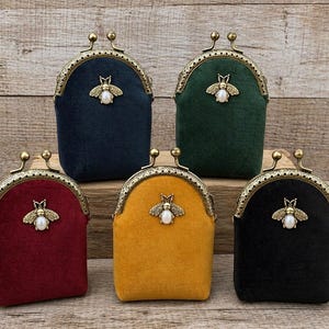 May include: Five velvet coin purses in navy, forest green, burgundy, mustard yellow, and black. Each purse features a brass-toned metal kiss-lock frame and a central bee-shaped embellishment with a white faux pearl body at the top center.