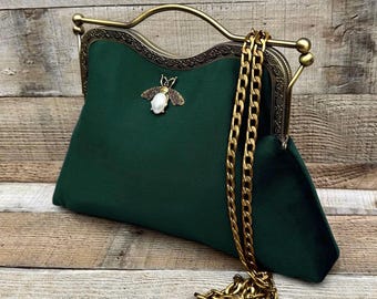 May include: A dark green handbag with a gold-colored chain strap and clasp. The bag features a decorative bee emblem with a pearl-like body. The clasp has an ornate design, and the bag is made of a soft, velvety material.