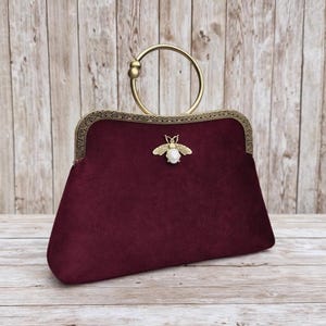 Puede incluir: Bolso de mano de terciopelo burdeos con asa y marco dorados. El bolso presenta un adorno decorativo en forma de abeja con un centro perlado. El asa tiene un diseño circular.