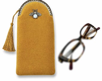 May include: Mustard yellow glasses case with a decorative bee emblem and a tassel. The case has a metal clasp and a soft, velvety texture. Tortoiseshell eyeglasses are slightly out of focus in the background.