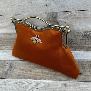 Puede incluir: Un bolso de mano de color óxido con un marco y asa de metal de tono bronce. El bolso presenta un emblema decorativo de abeja con una piedra ojo de tigre. El marco tiene un borde cosido decorativo.