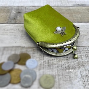 May include: A lime green velvet coin purse with a decorative butterfly clasp. The purse has a vintage-style metal frame and a pearl accent. Several coins are scattered in the foreground.