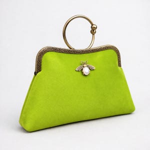May include: A lime green clutch bag with a gold-coloured handle and clasp. The clasp has an ornate design. A gold-coloured bee-shaped brooch with a pearl is attached to the front. The bag is made of a soft, velvety material.