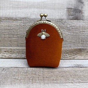 May include: A rust-colored suede coin purse featuring a brass-toned metal kiss-lock frame and a central bee-shaped embellishment with a white oval stone inlay. The purse has a rounded top and a structured, compact, vertical rectangular shape.