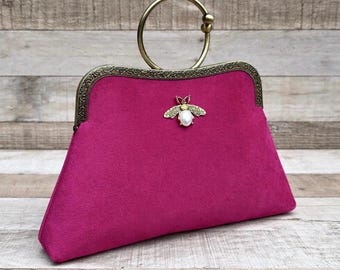 Op de afbeelding: Fuchsia clutch met een goudkleurige handgreep en sluiting. De tas heeft een decoratieve bijenversiering met een parelaccent. Het frame heeft een sierlijk ontwerp in vintage-stijl.